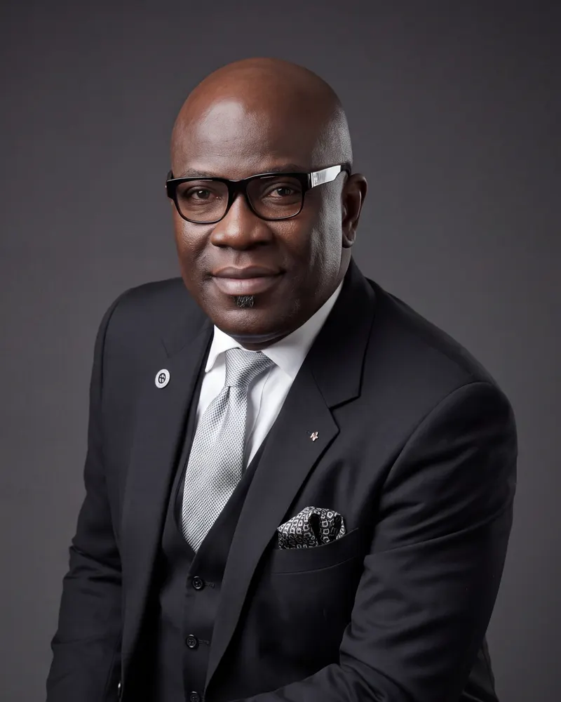 David Amodu in a dark three-piece suit and silver tie, against a charcoal studio backdrop
