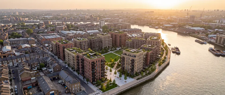 Aerial view at golden hour over a riverside regeneration area: new brick apartment buildings around a park, cranes on the horizon