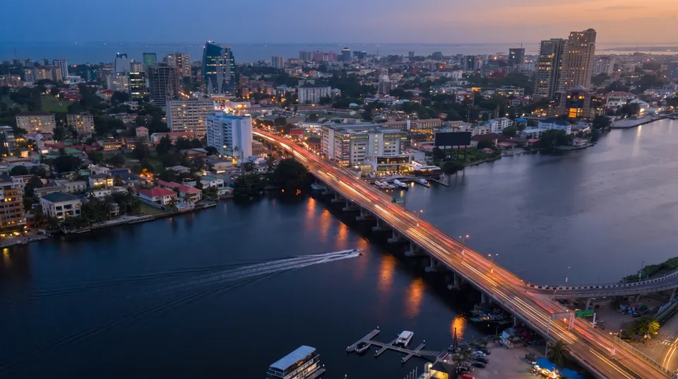 Lagos at dusk: the Falomo Bridge crossing the Five Cowries Creek, with red roofs, palms and the city's towers beyond