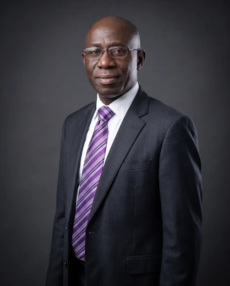 Dr Mustapha Ojo in a charcoal suit and purple striped tie, against a charcoal studio backdrop